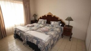 A double bed with a patterned bedspread, two bedside tables with lamps, and a window with curtains. - Bluewater Guesthouse (Gqeberha (Port Elizabeth))