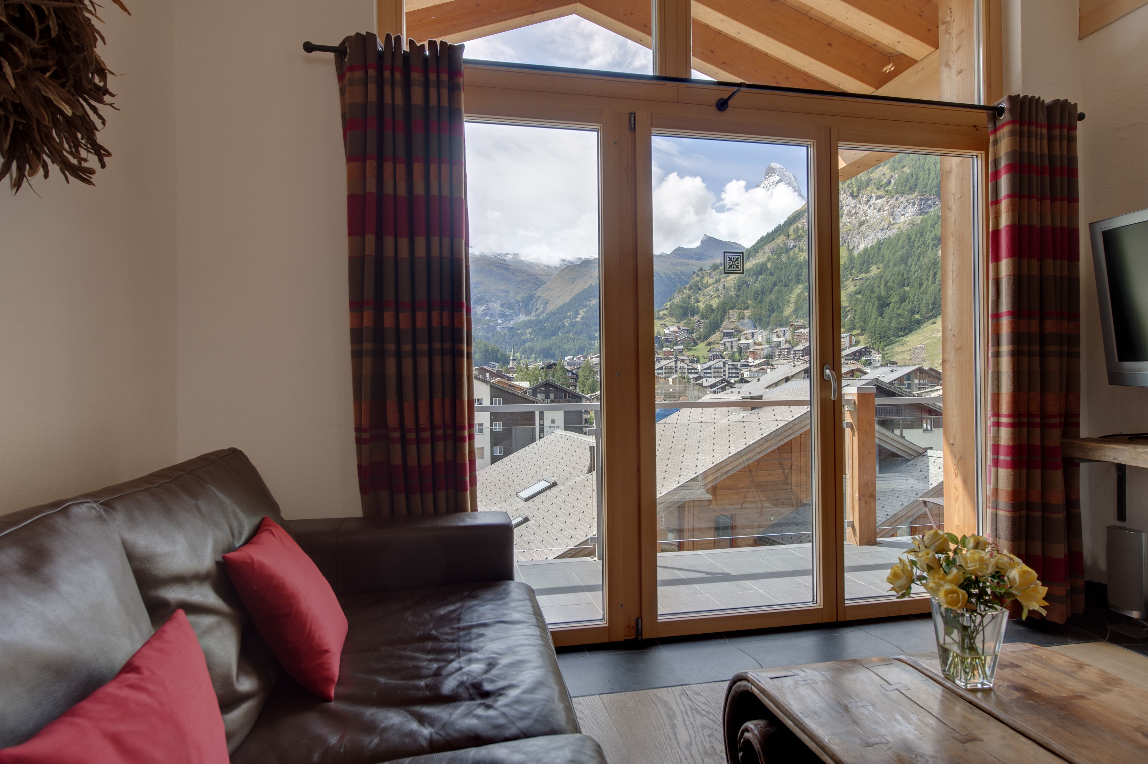 A living room with a leather sofa, a coffee table, and a view of a mountainous village through large glass doors.