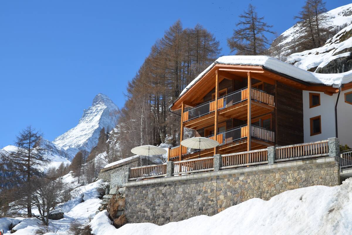 A chalet with a stone foundation, wooden balconies, and a view of a snow-covered mountain.