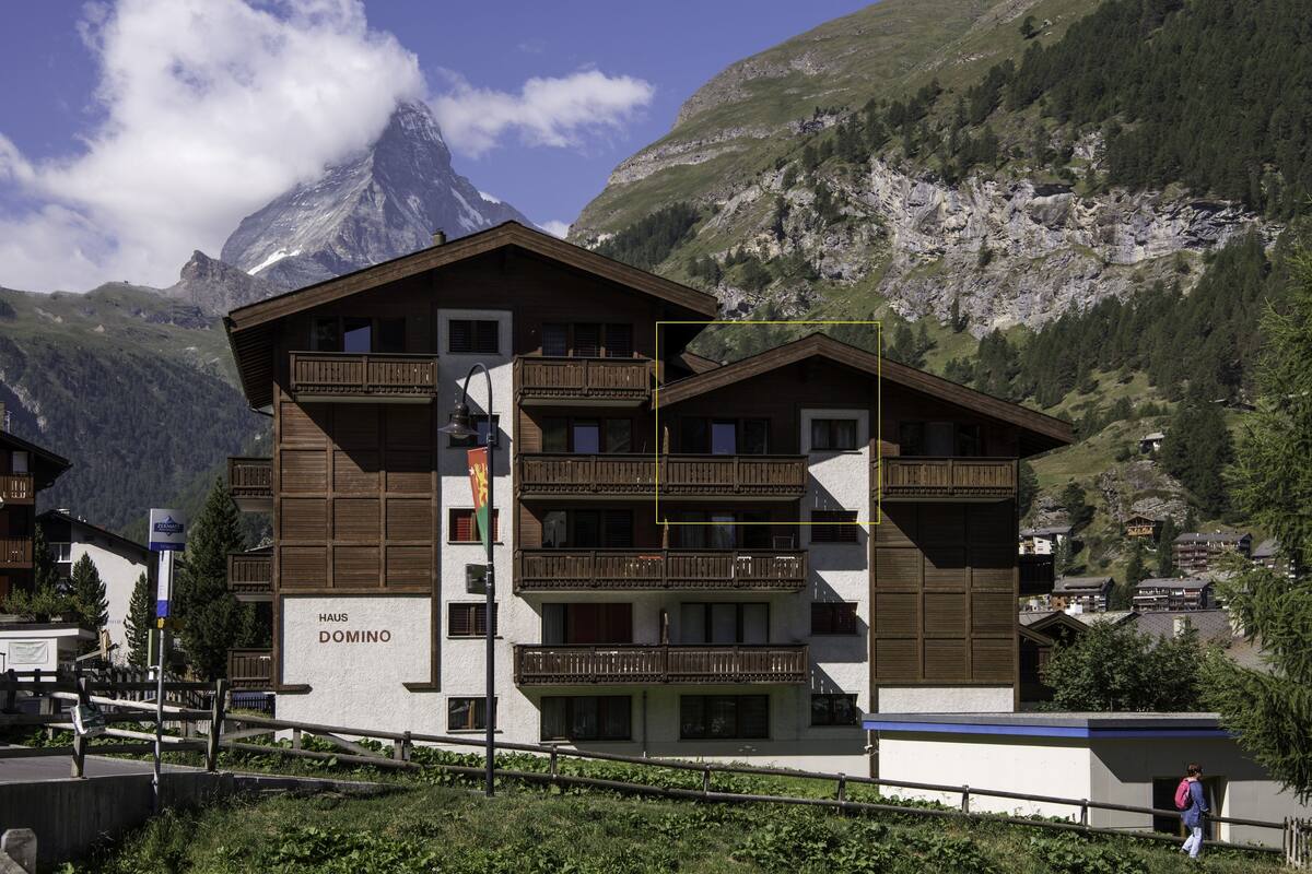 A multi-story building with balconies, labeled "HAUS DOMINO," set against a mountainous backdrop.