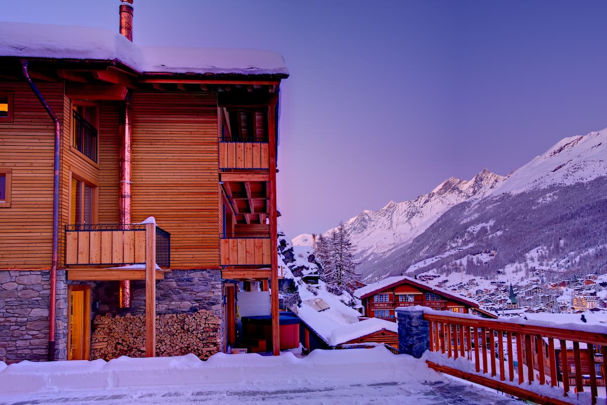 A wooden lodge with a stone foundation, wooden balconies, and a snow-covered landscape with mountains in the background.