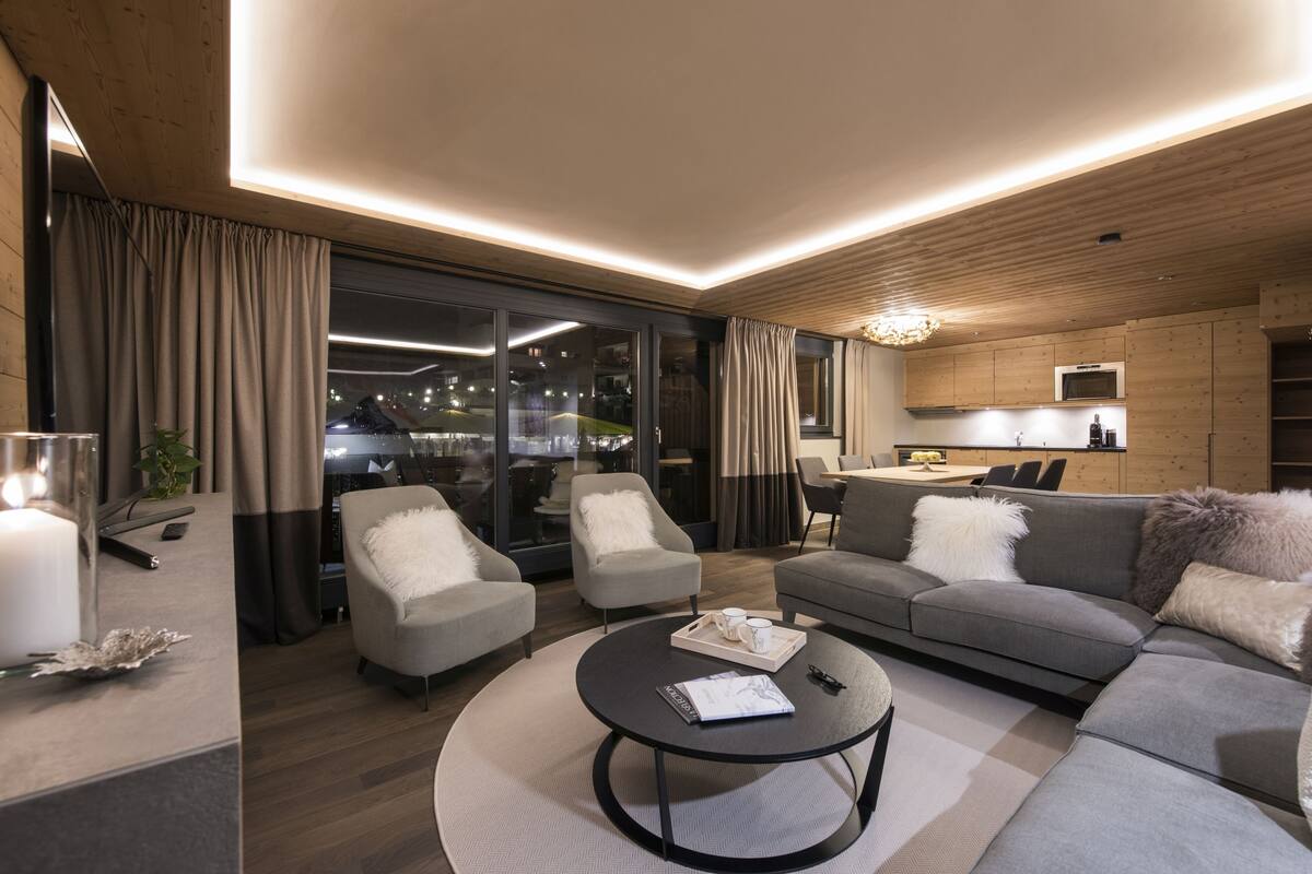 A modern living room with a sofa, armchairs, a coffee table, and a dining area with a kitchen in the background.
