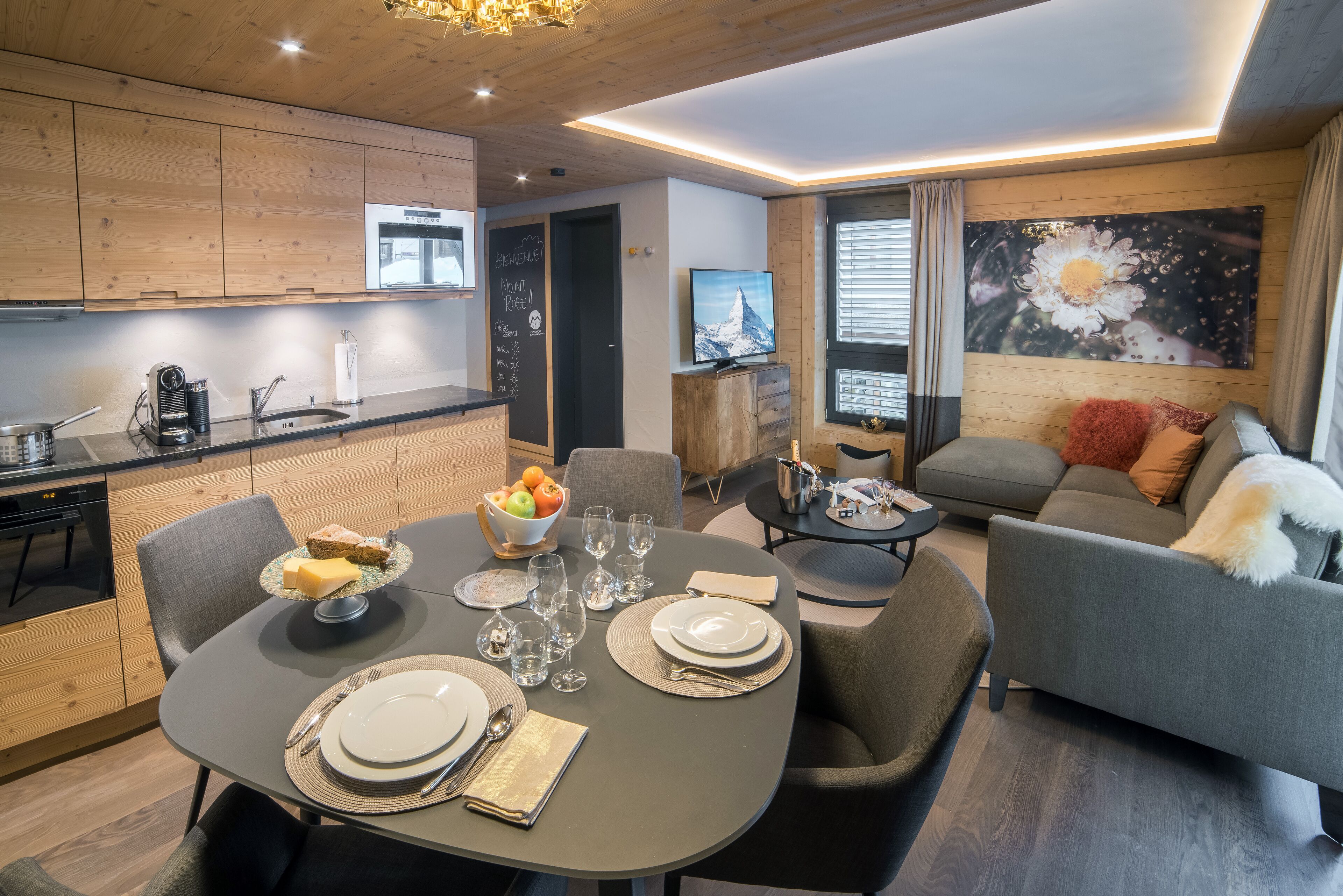 A modern living room with a grey sofa, a dining table set with plates and glasses, and a kitchen area with wooden cabinets and a blackboard.