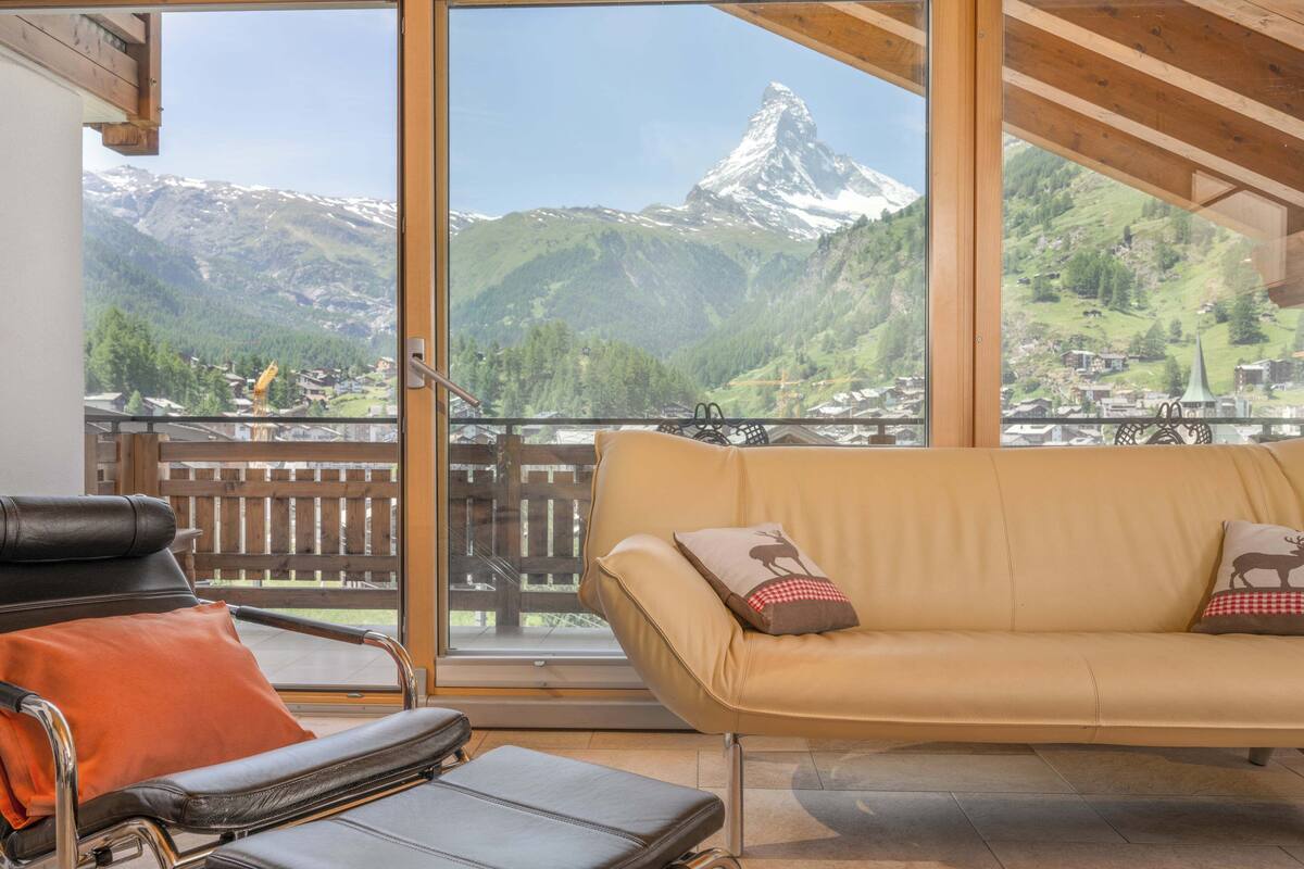A room with a sofa and a leather chair facing a large window with a mountain view.