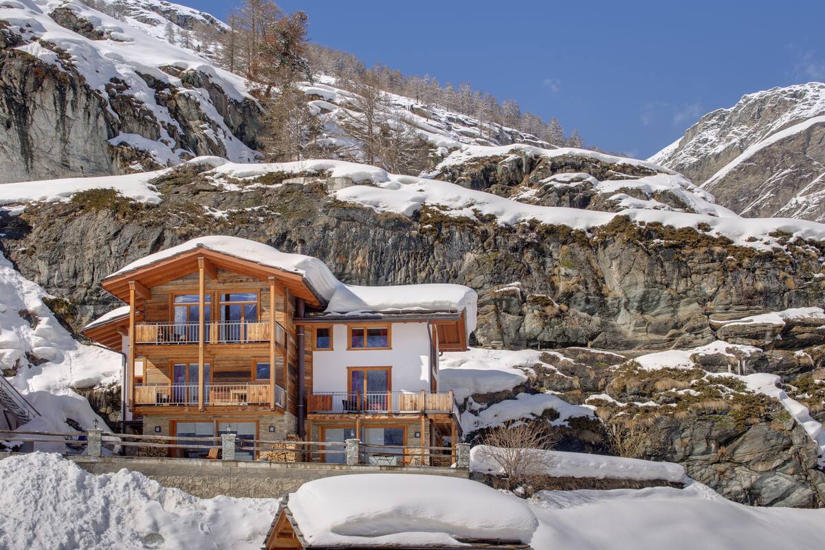 A multi-story wooden chalet with balconies, set against a mountainous backdrop with snow.