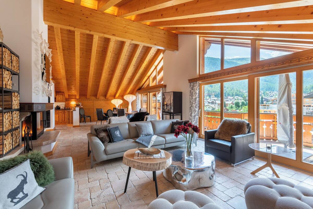 A spacious living area with a high wooden ceiling, a fireplace, a sofa set, and large windows offering a view of the mountains.