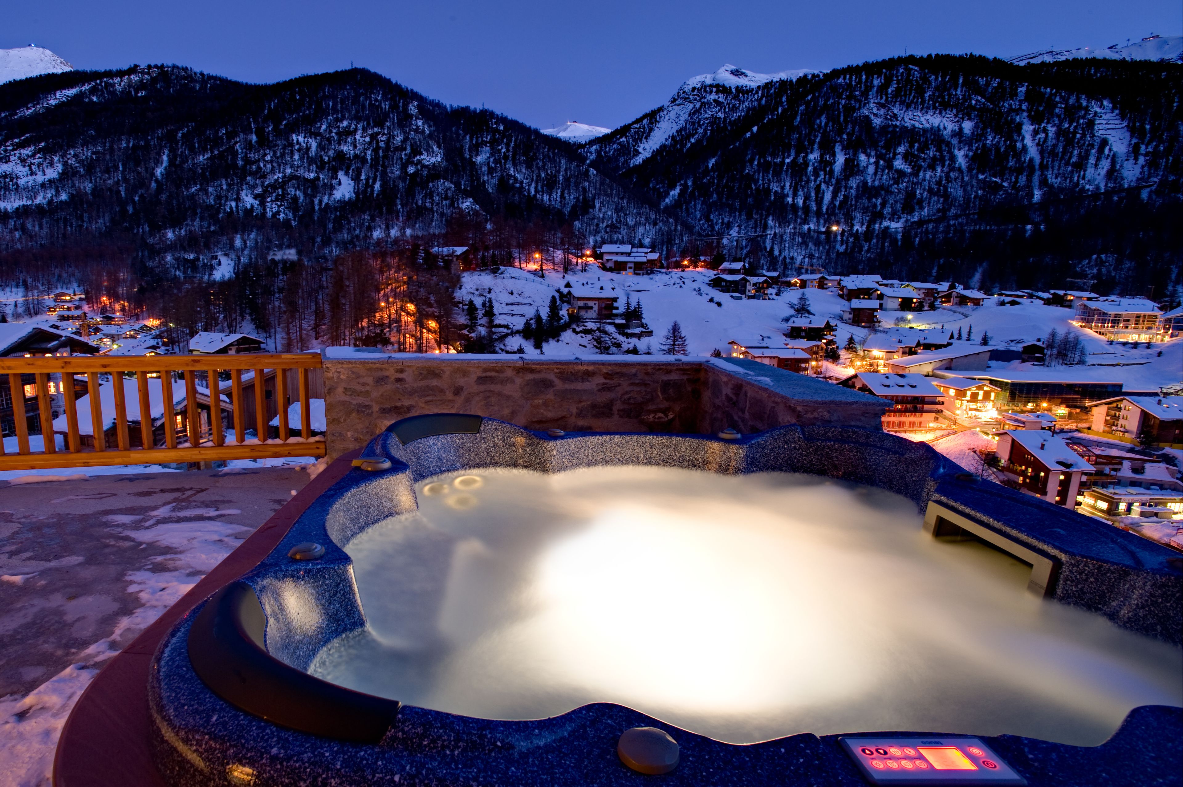 A hot tub with a digital timer, surrounded by a wooden deck, overlooking a snow-covered village with buildings and mountains in the background.