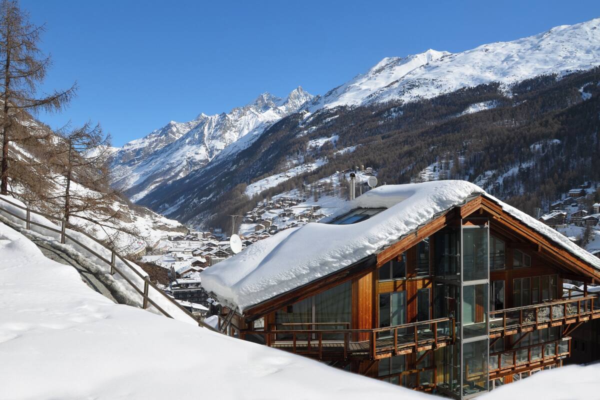 A wooden cabin with large windows, surrounded by snow-covered mountains and a clear sky.