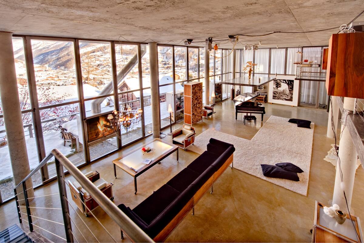 A spacious living area with large windows, a fireplace, a piano, and a view of a snowy landscape.