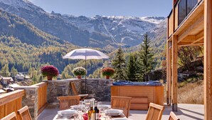 Un área de comedor al aire libre con una mesa dispuesta para seis personas, rodeada de sillas de madera, con vista a un paisaje montañoso y cielo despejado.