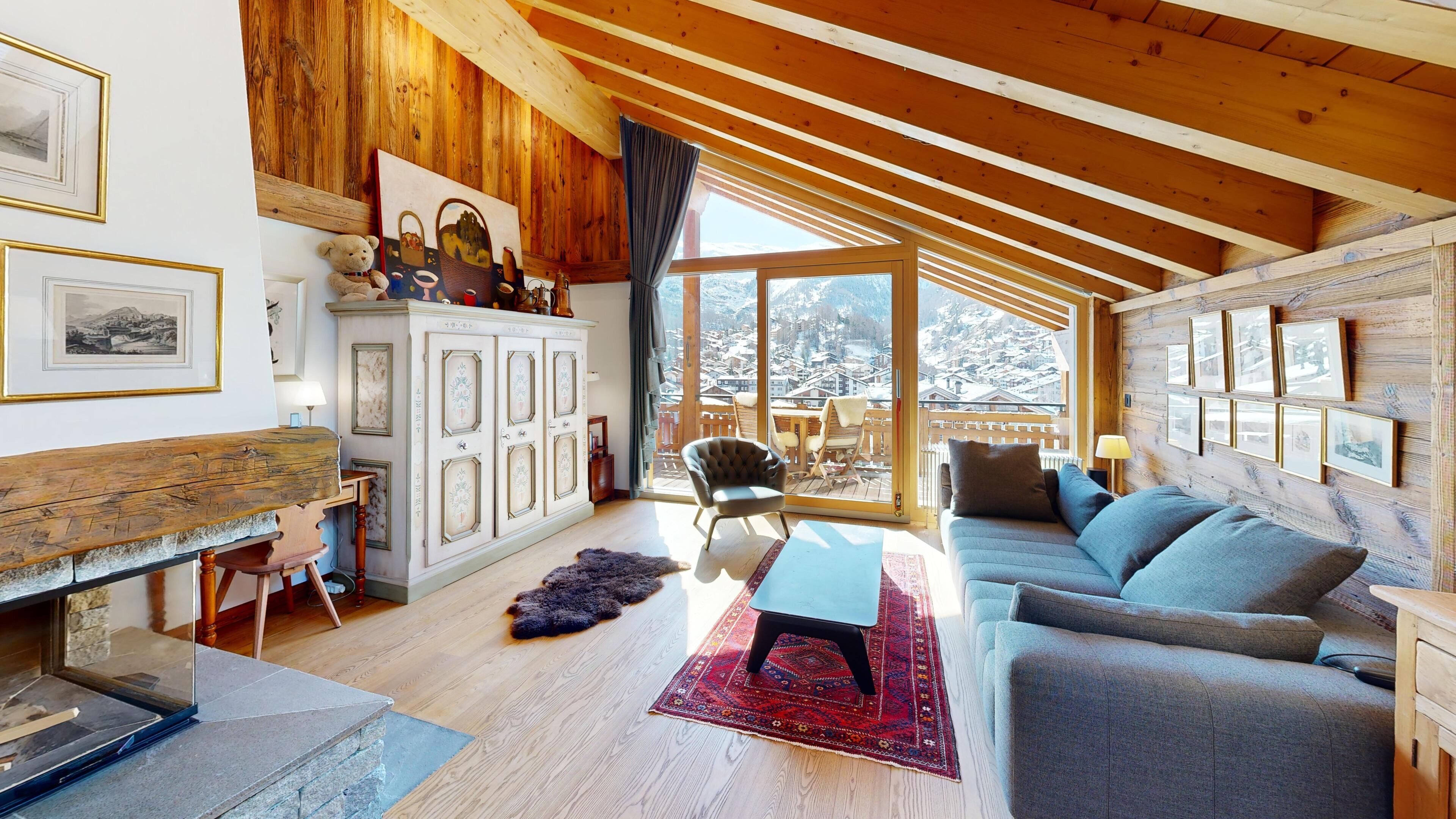 Un salon chaleureux avec un plafond en bois, un canapé, un fauteuil, un meuble contenant des photos encadrées et une vue sur des montagnes enneigées, visible à travers de grandes fenêtres.