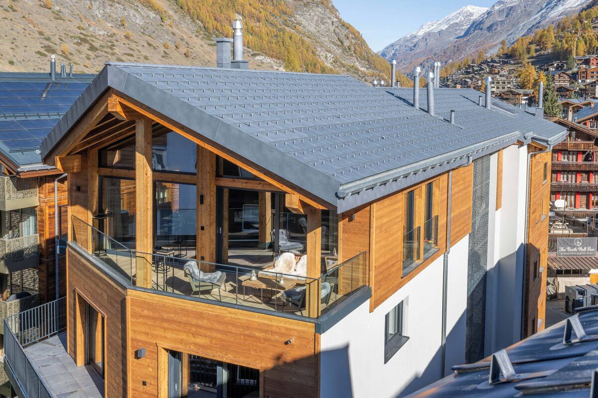 A modern wooden building with a balcony, large windows, and a mountain backdrop.