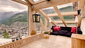Una habitación con techos altos y detalles de madera, un sofá negro con cojines rosados, una mesita y vistas a un pueblo y montañas a través de amplios ventanales.