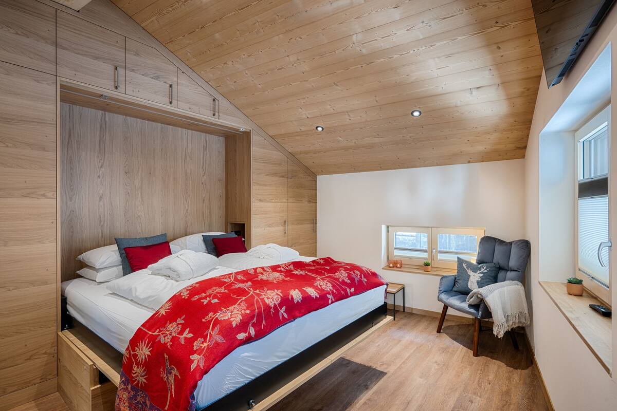 A bedroom with a wooden ceiling, a bed with a red floral bedspread, a chair with a deer pillow, and a window with blinds.