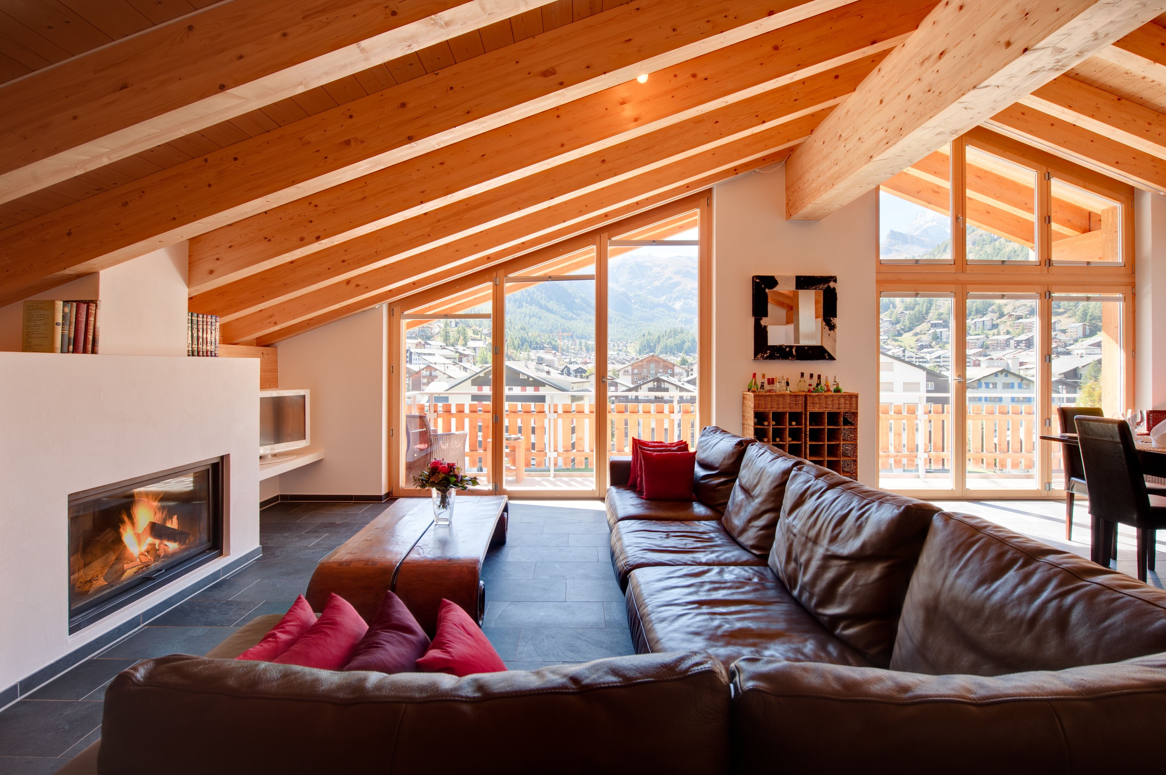 A spacious living room with a high ceiling, a fireplace, a leather sofa, and large windows offering a view of a mountainous landscape.
