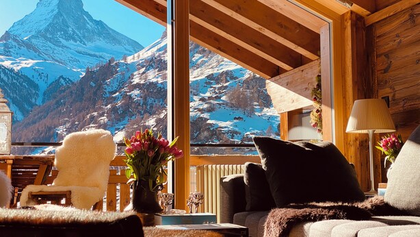 Habitación acogedora con un ventanal grande que ofrece vistas a una montaña. Hay un sillón cómodo, un sofá y una mesa con un jarrón de flores.