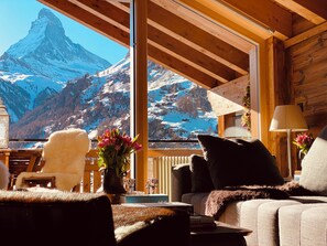 A cozy room with a large window offering a view of a mountain. There is a plush chair, a sofa, and a table with a vase of flowers. - Luxury Chalets & Apartments by MX Zermatt (Zermatt)