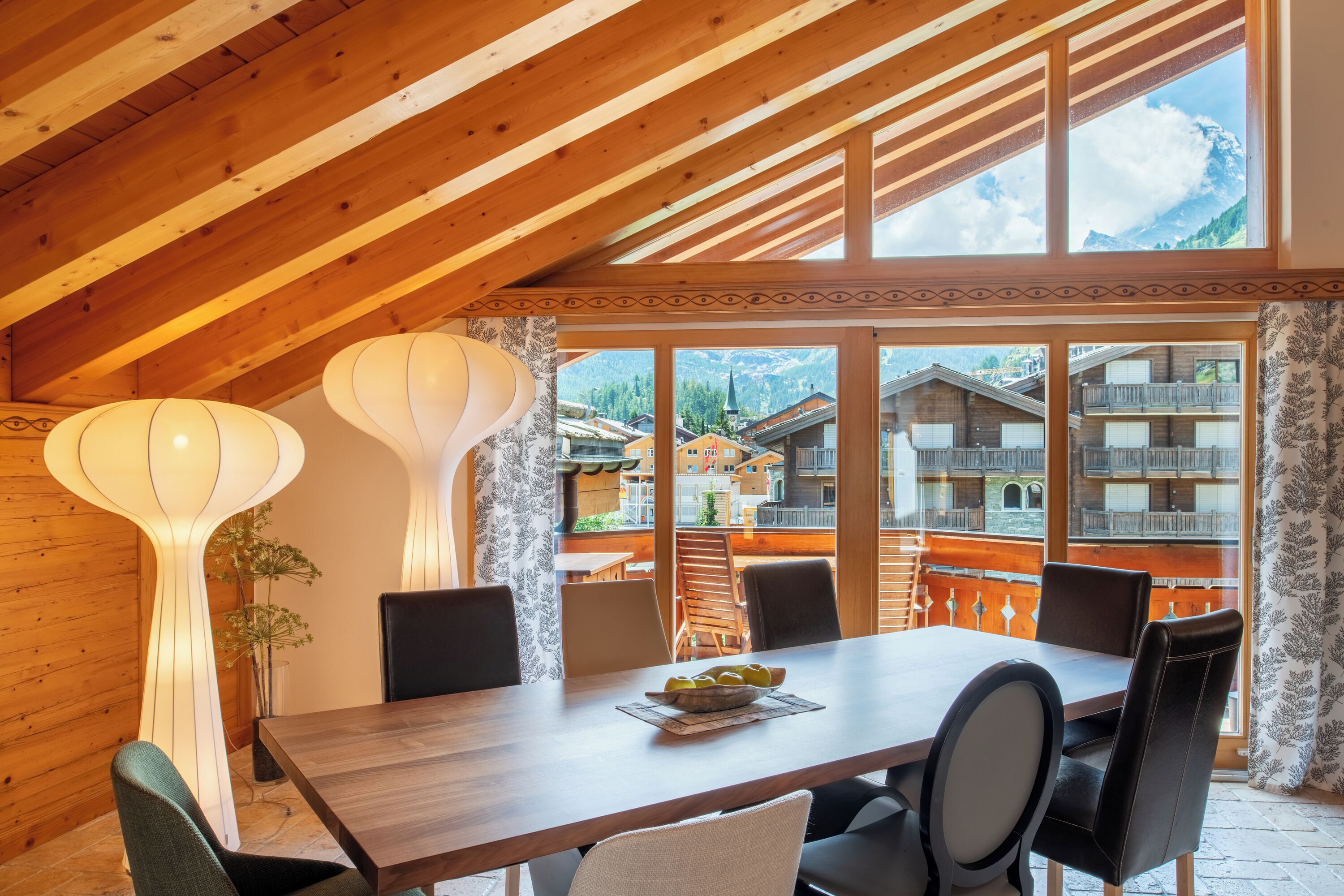Un comedor con una mesa y sillas de madera, ventanas amplias con vistas a la montaña y lámparas decorativas en la pared.