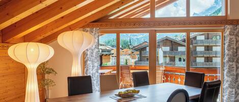 Un comedor con una mesa y sillas de madera, ventanas amplias con vistas a la montaña y lámparas decorativas en la pared.