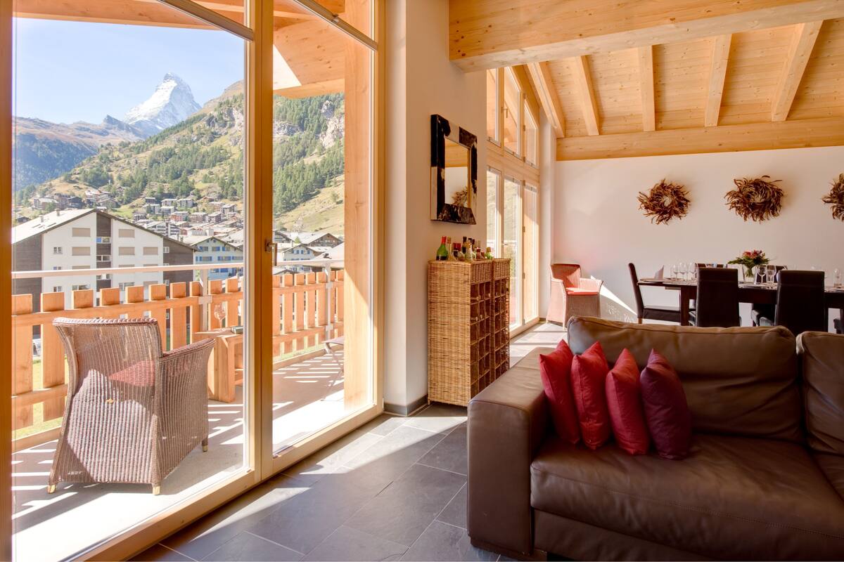 A balcony with a wicker chair, a brown sofa with red cushions, a wicker side table, and a view of a mountain.