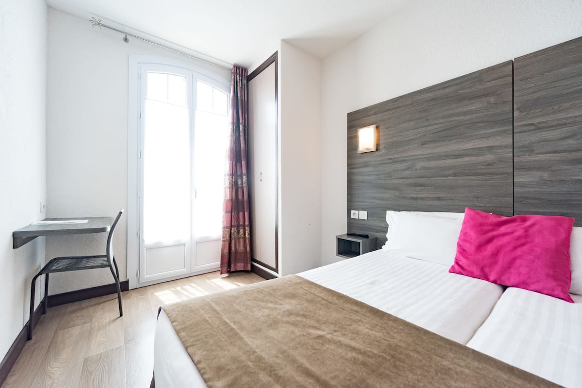A hotel room with a bed, a desk, a chair, a nightstand, and a window with curtains.