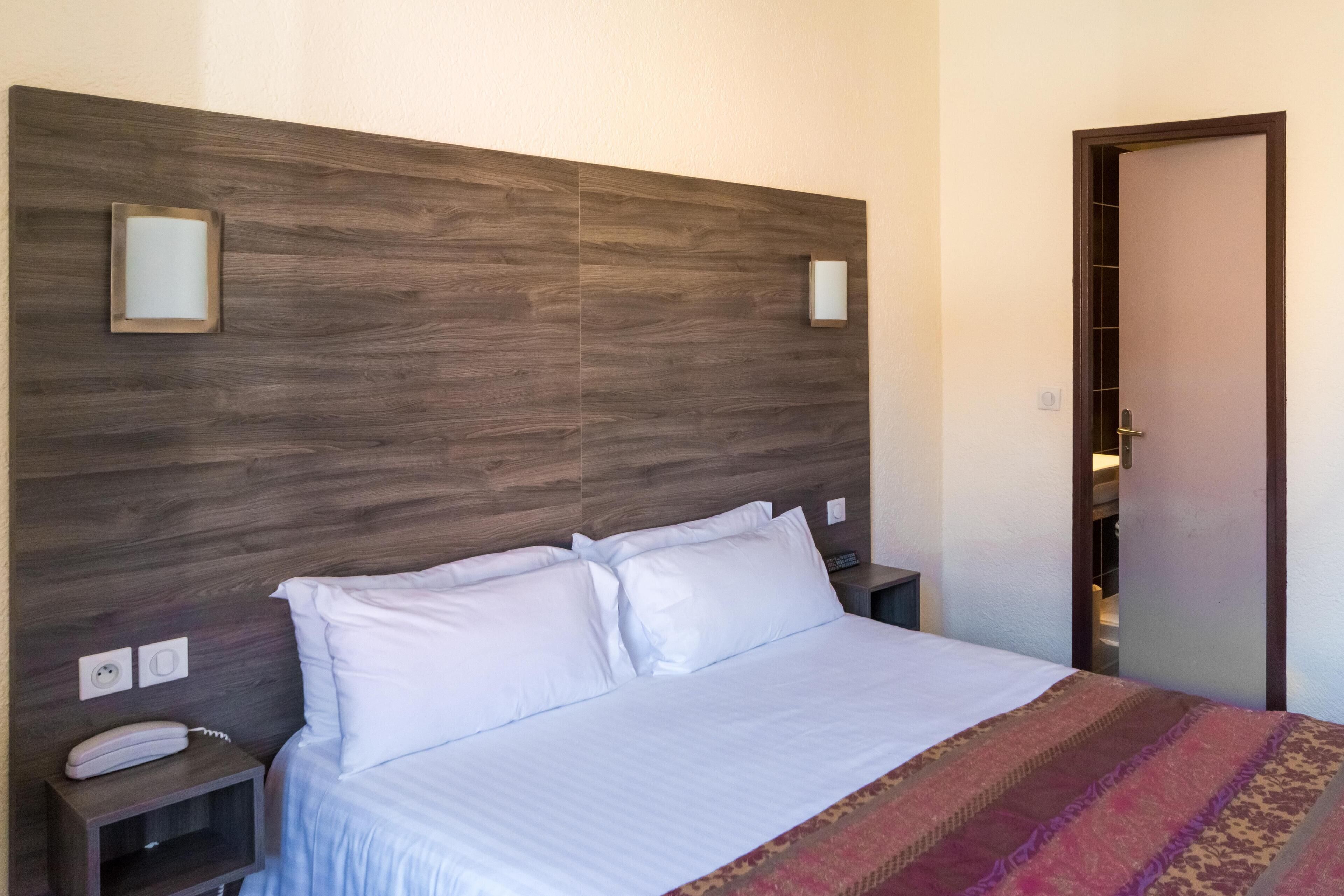 Una cama bien tendida con sábanas blancas, cabecero de madera, mesitas de noche con un teléfono y una lámpara, y una manta decorativa.