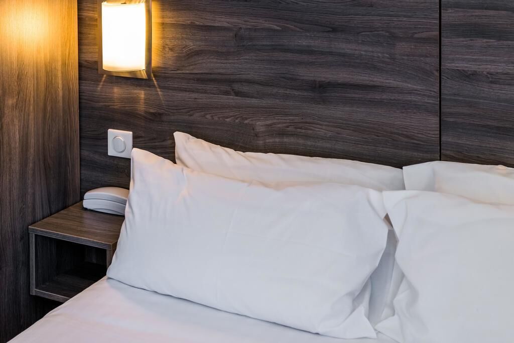 Ein ordentlich bezogenes Bett mit weißen Leinen, ein Nachttisch mit Lampe und eine Wandleuchte.