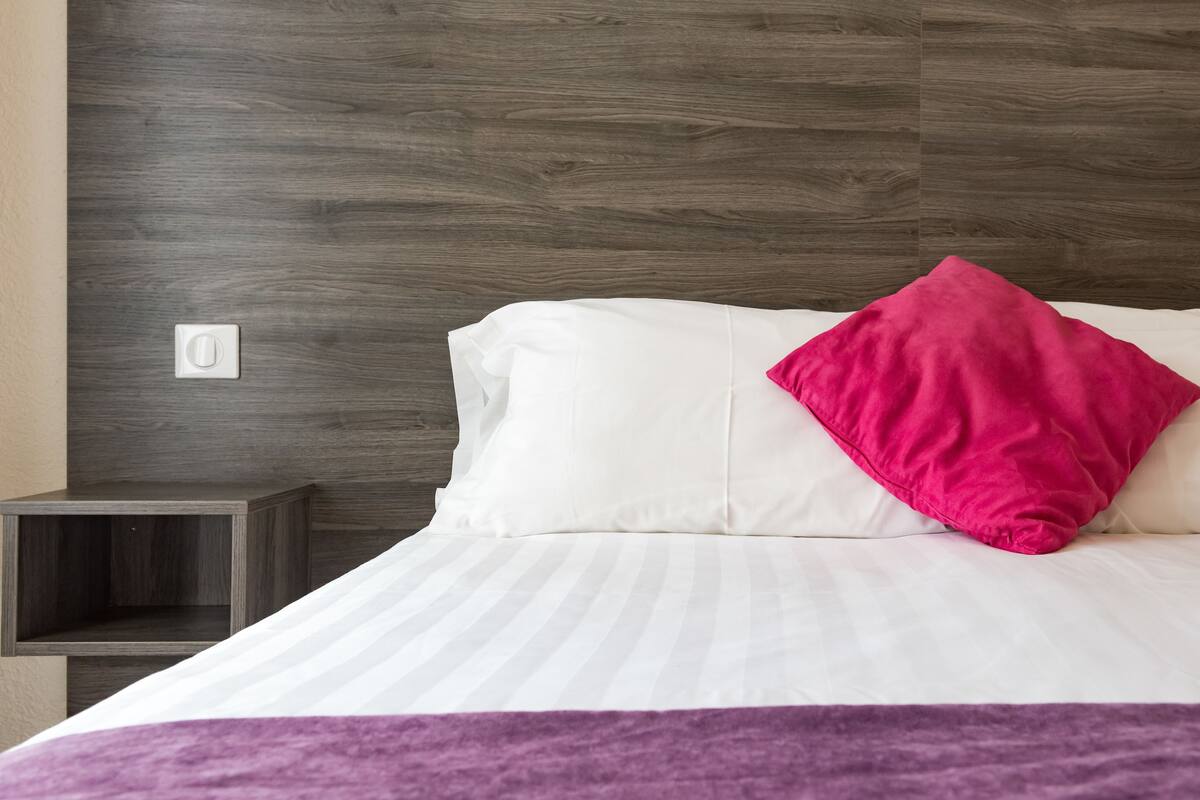 A neatly made bed with white and pink pillows, a purple blanket, and a dark wooden headboard.