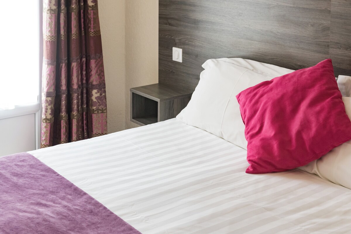 Una cama bien hecha con una colcha blanca y morada, una sola almohada blanca y una almohada rosa brillante.