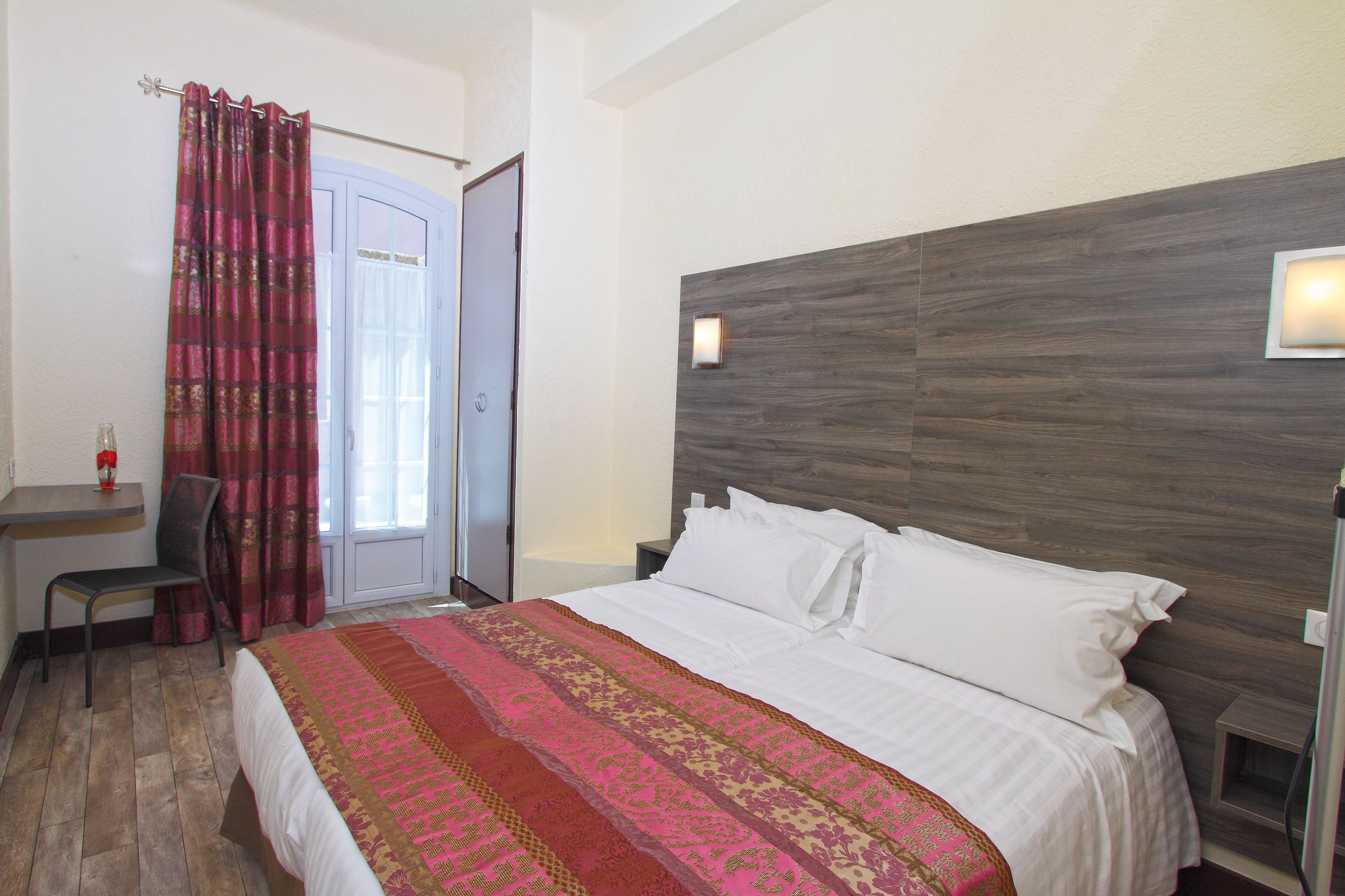 Ein Hotelzimmer mit hölzernem Kopfteil, einem Bett mit weißen Leinen, einer gemusterten Tagesdecke, einem Schreibtisch mit Stuhl und einem Fenster mit Vorhängen.