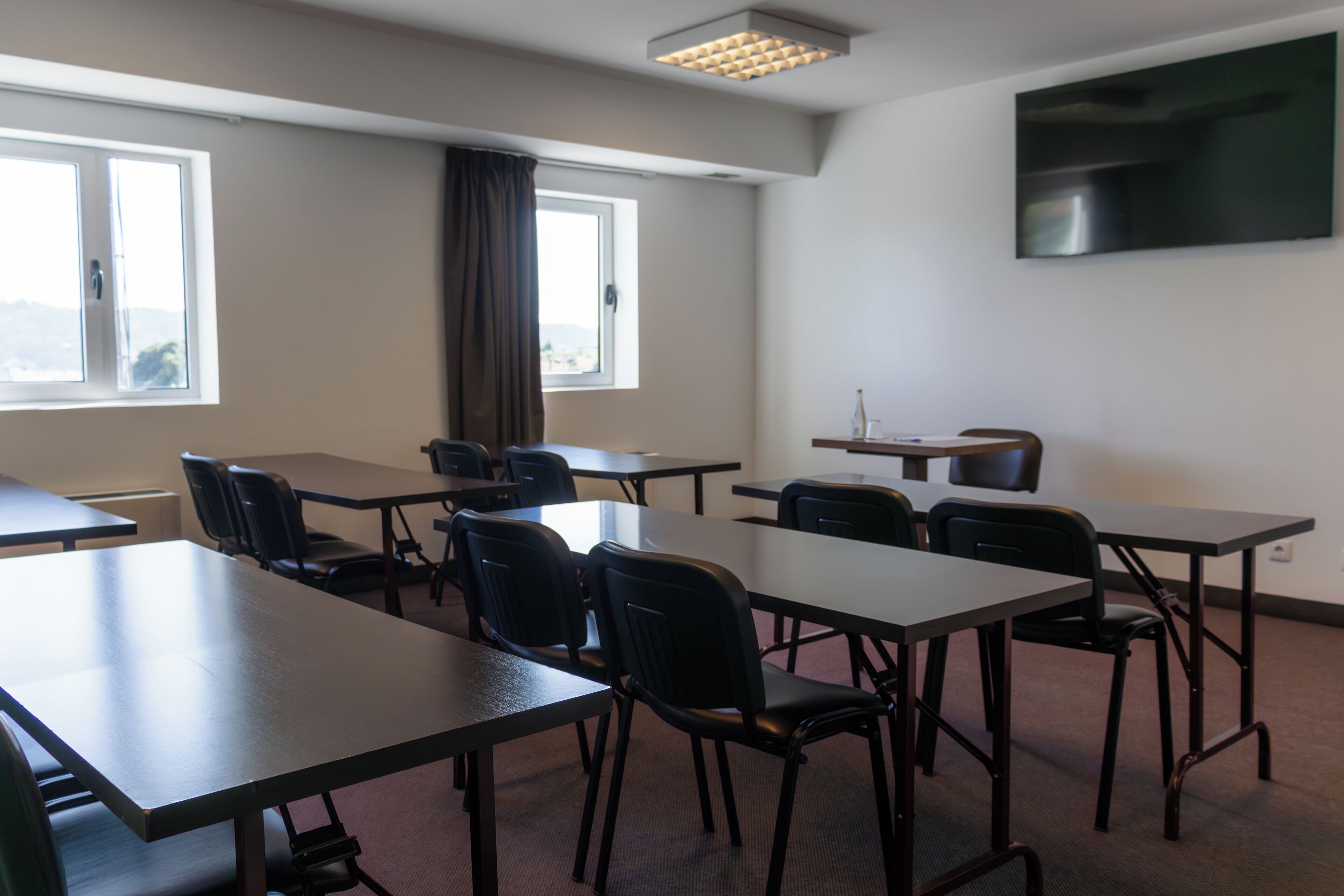 A conference room with tables and chairs, a large window, a flat-screen TV on the wall, and a view of the outdoors.