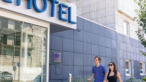 Entrada de un hotel con puerta de vidrio, un cartel que dice 'HOTEL' y una persona con maleta.