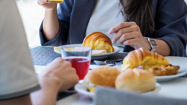 Una persona sentada a una mesa con un croissant, un vaso de jugo de naranja y una bebida roja.