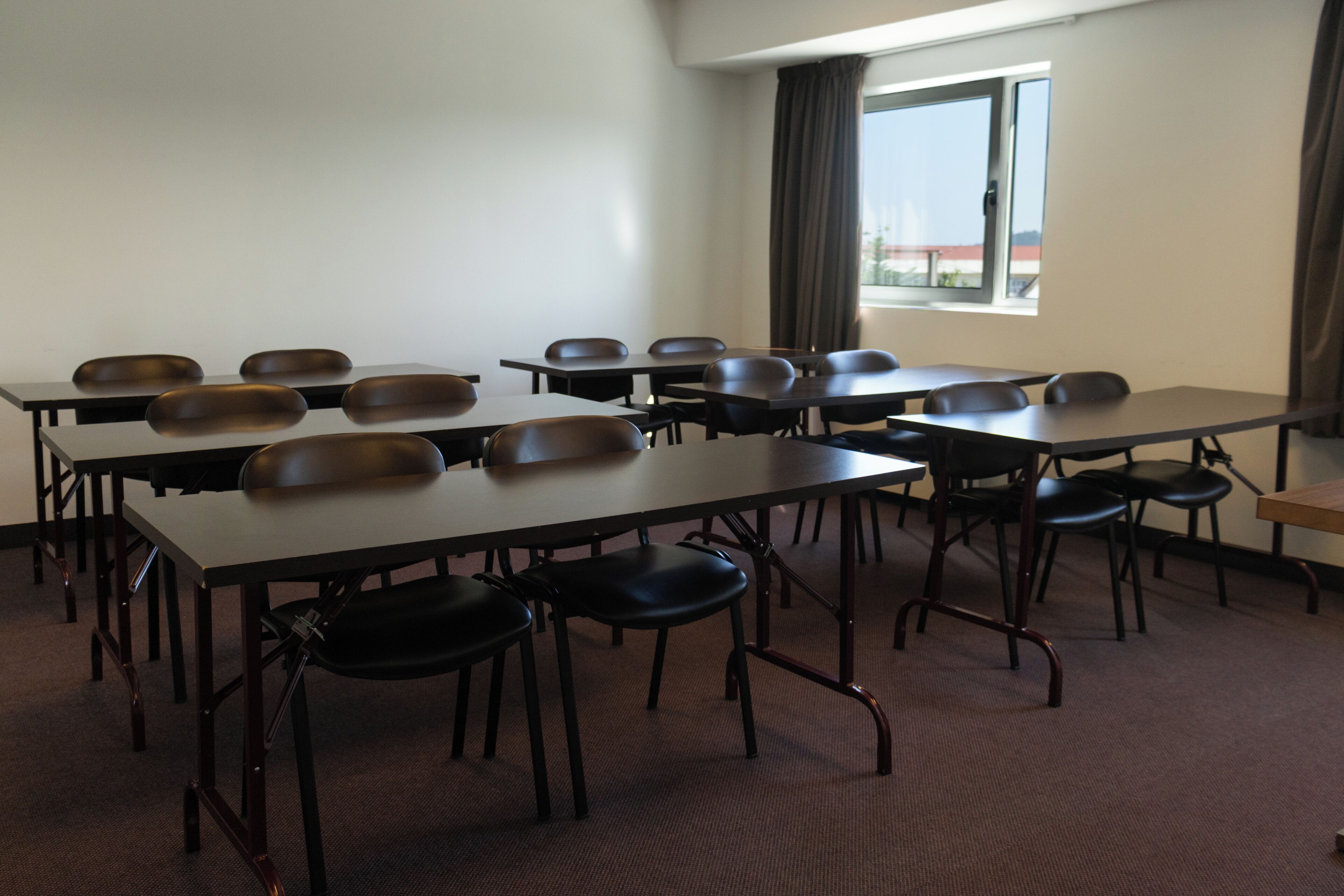 A conference room with multiple tables and chairs, a large window with curtains, and a carpeted floor.