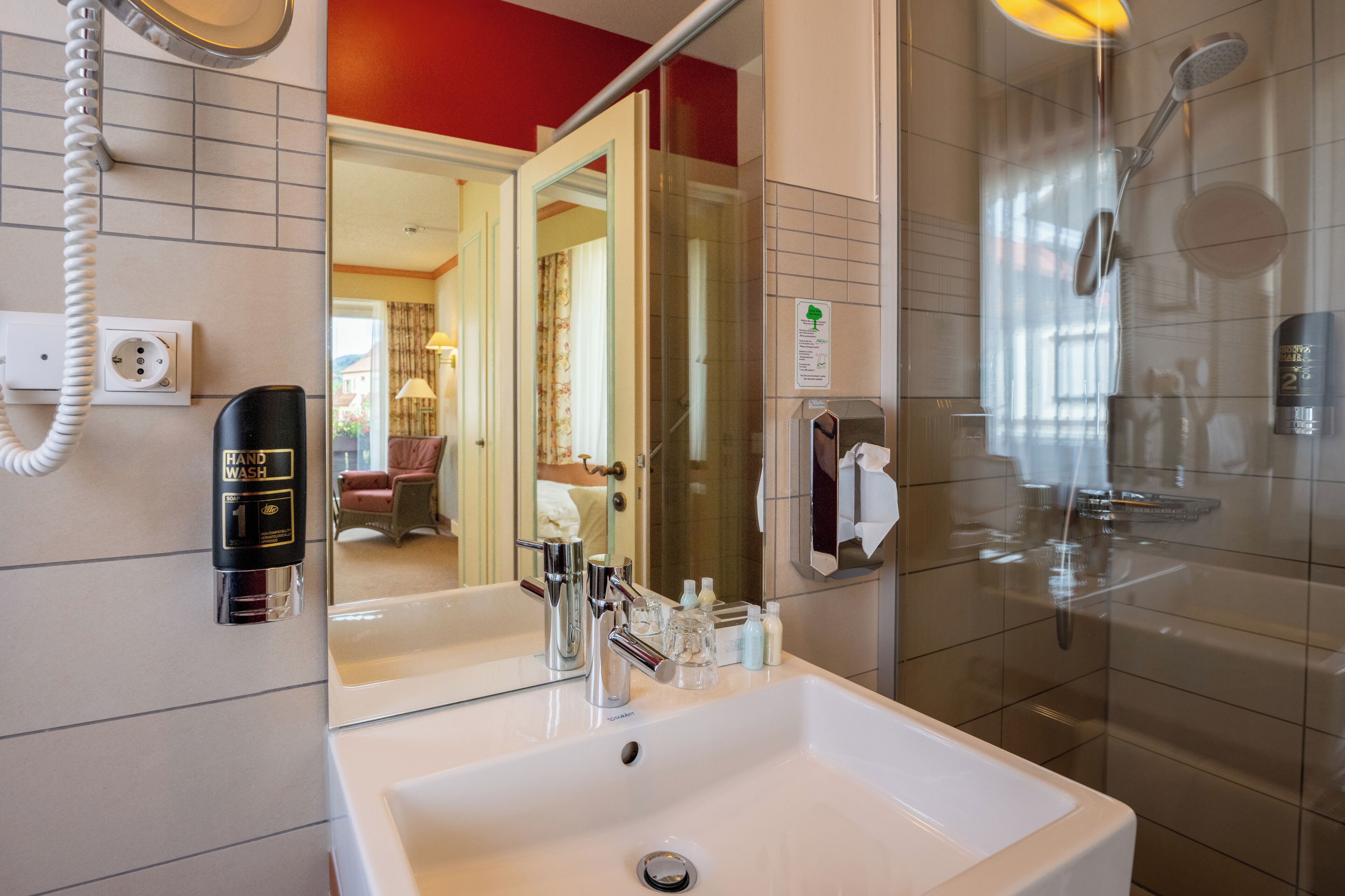 A hotel bathroom with a sink, mirror, hand wash station, and a shower.