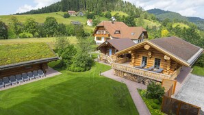 Eine Blockhütte mit Balkon, Rasen und Blick auf umliegende Häuser und Hügel.