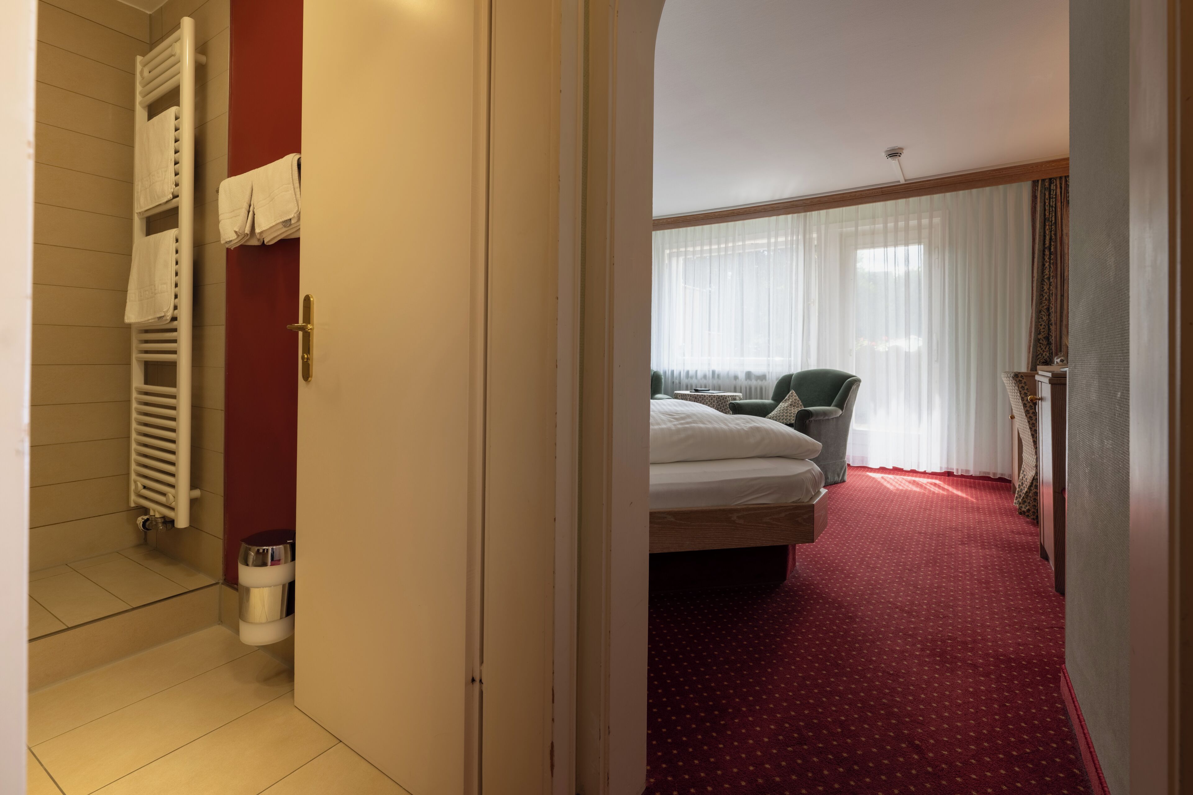 A hotel room with a bed, a chair, a radiator, and a window with curtains.