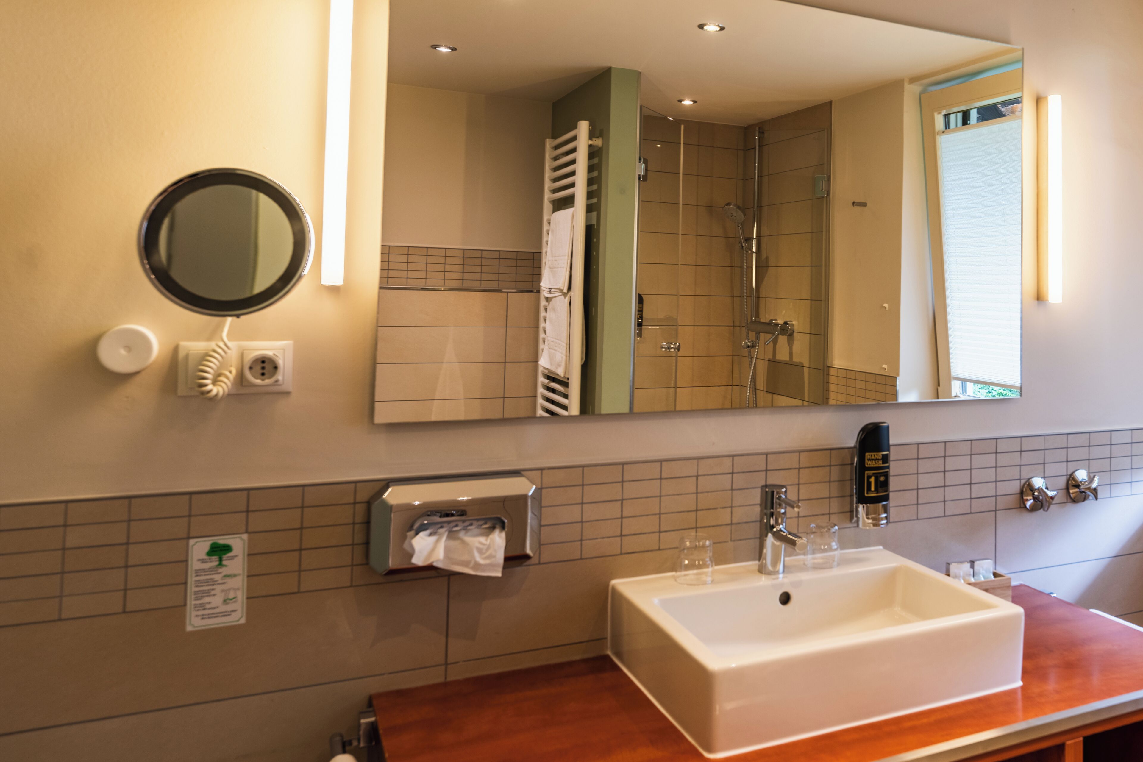 Un baño con lavamanos, espejo, dispensador de toallas y una ducha visible en el reflejo.