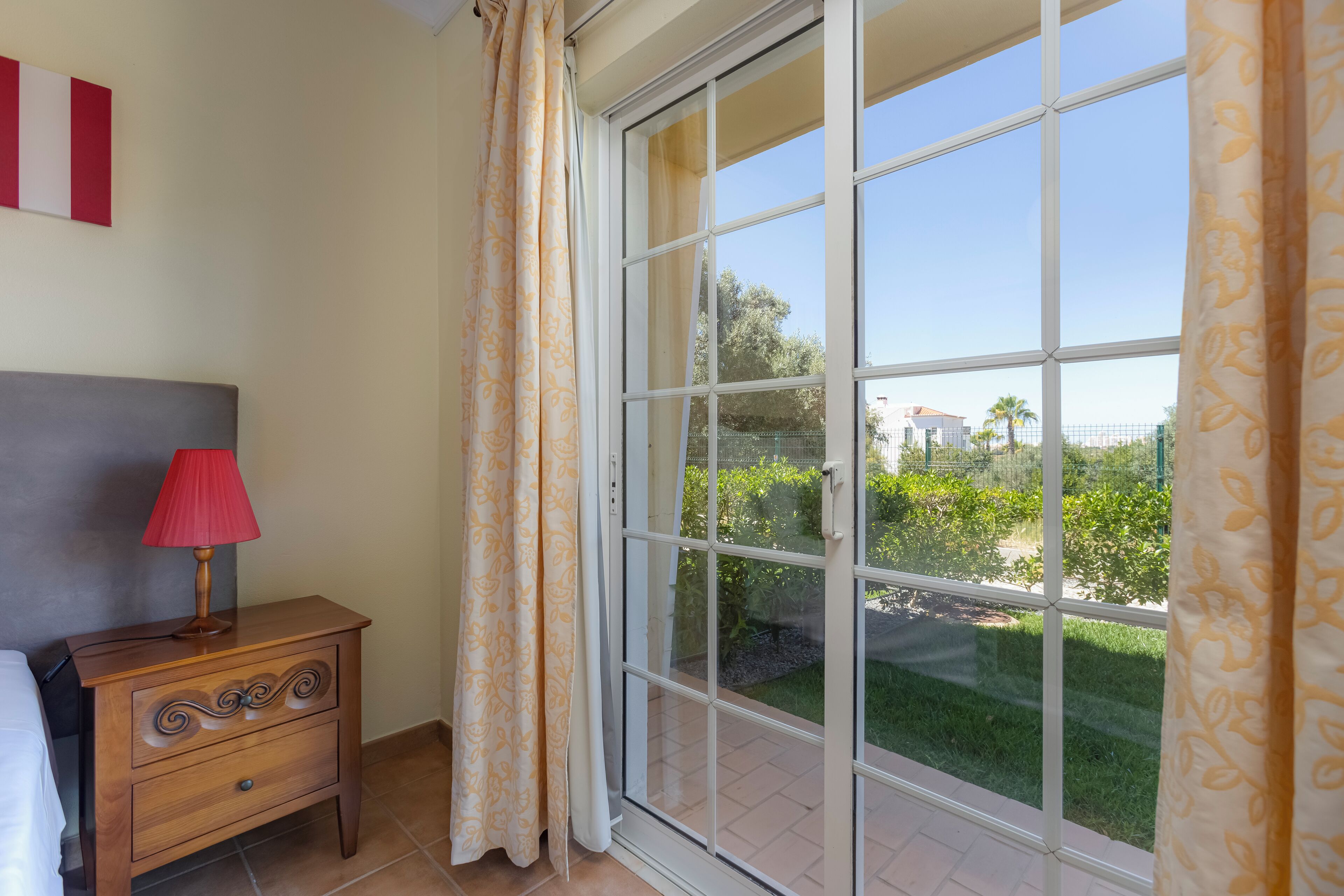 A bedroom with a nightstand, a lamp, a window with curtains, and a view of greenery outside.