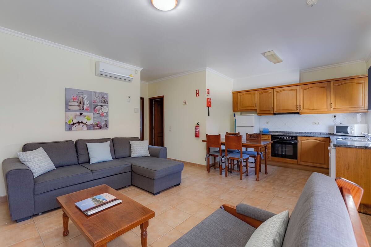 A living room with a grey sectional sofa, a wooden coffee table, and a dining area with wooden chairs and a table. The kitchen has wooden cabinets and a microwave.