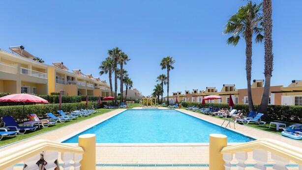 A pool area with lounge chairs and umbrellas, surrounded by palm trees and hotel buildings. - Villas Barrocal (Silves)