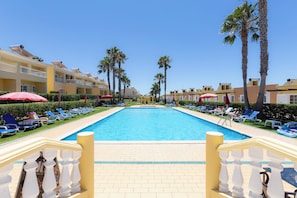 Zona de piscina con sillones de descanso y sombrillas, rodeada de palmeras y edificios de hotel.