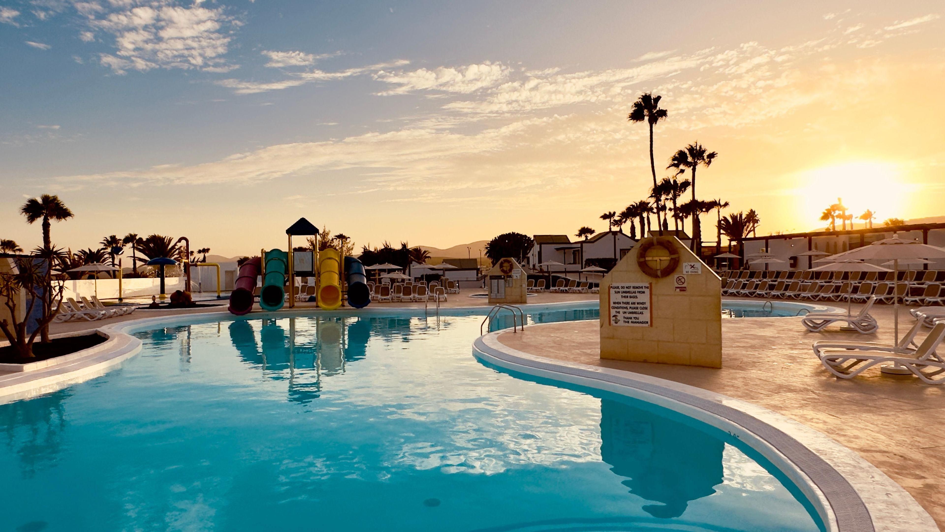 Una piscina con una torre de salvavidas, sillas de descanso y palmeras.