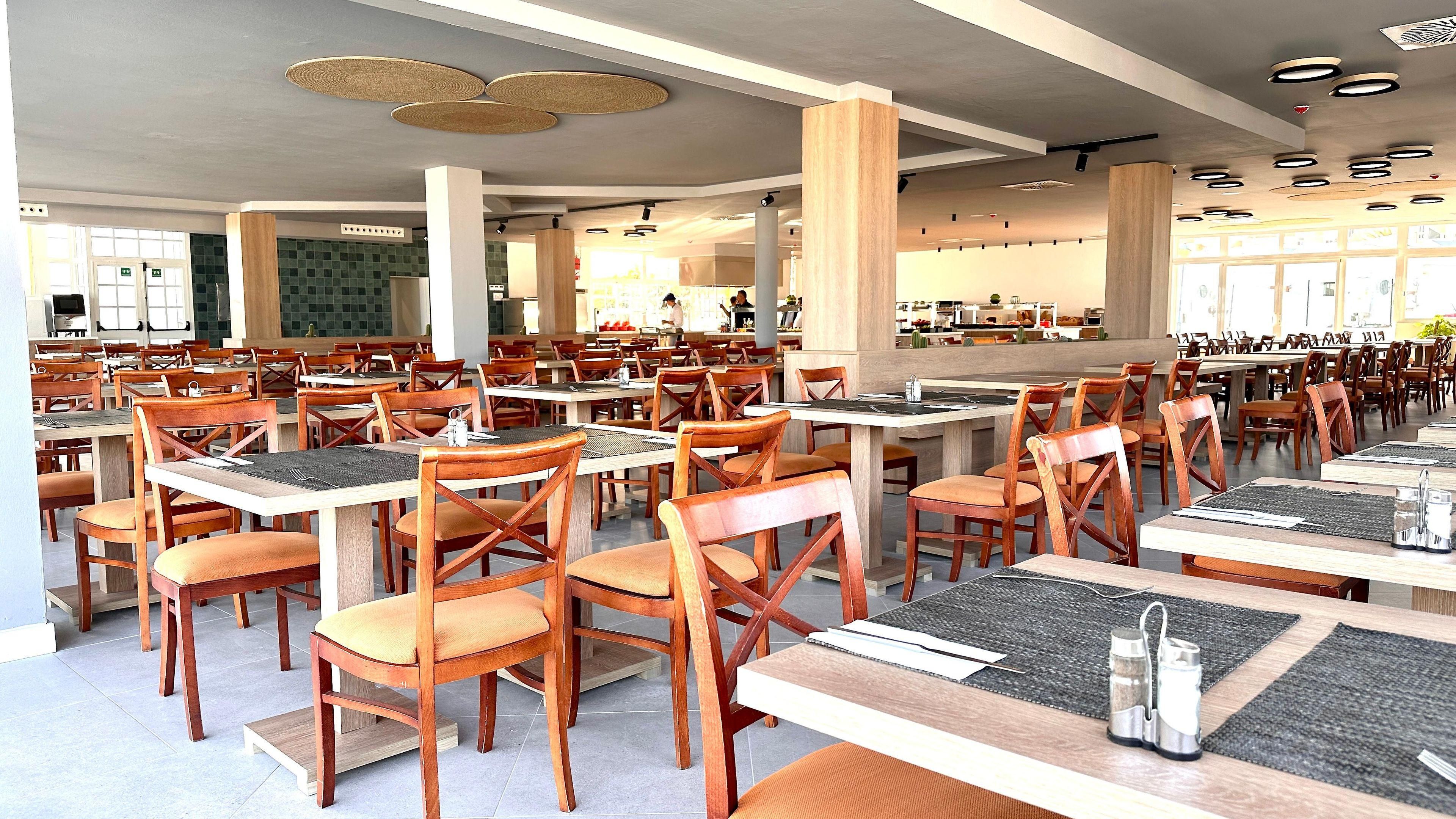 A spacious dining area with multiple tables and chairs, a kitchen in the background, and a modern ceiling design.