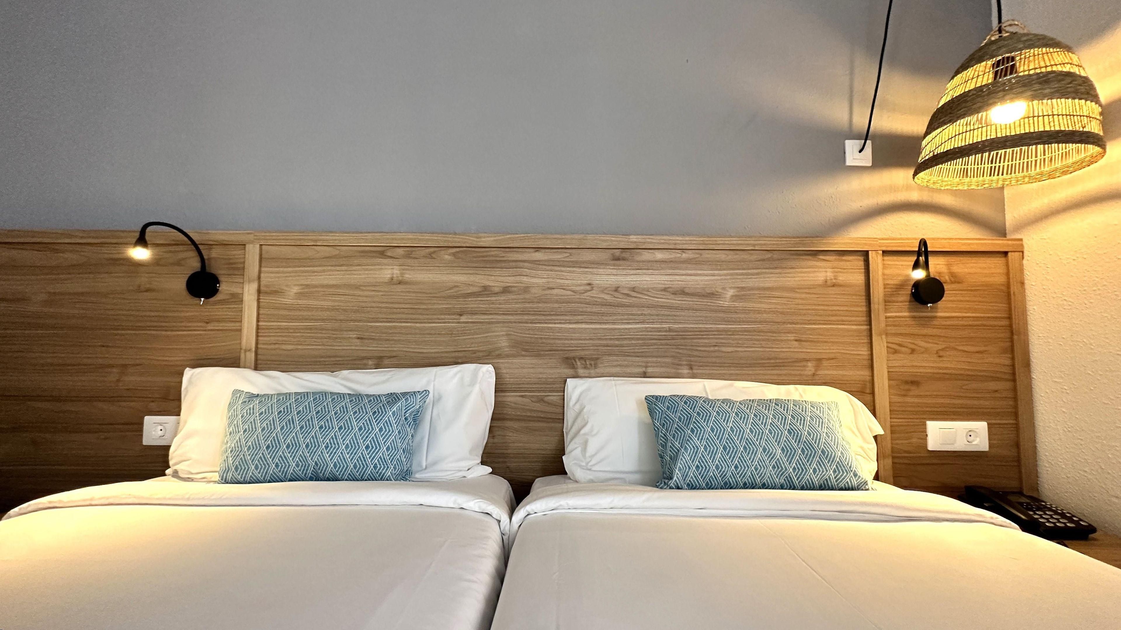 Una cama doble con ropa de cama blanca y almohadas estampadas, un cabecero de madera y dos lámparas fijadas a la pared.