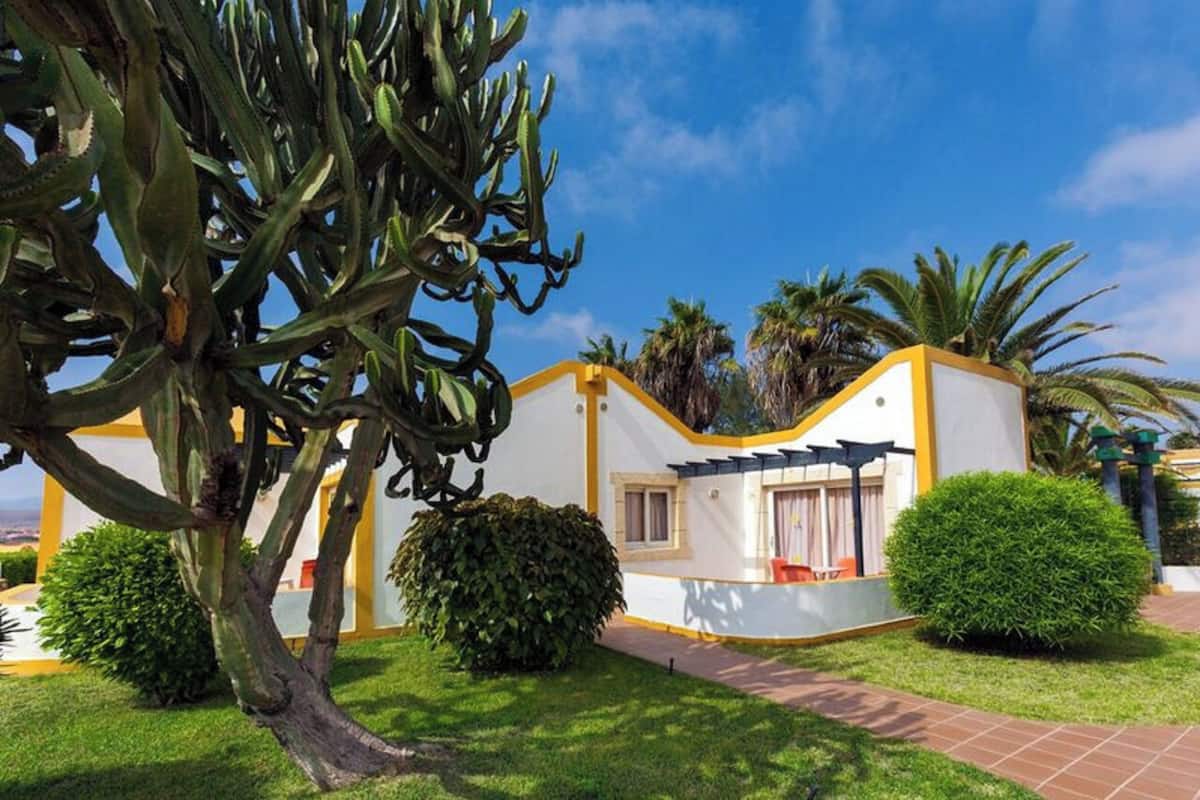 Una casa moderna con fachada blanca y detalles amarillos, rodeada de vegetación exuberante y palmeras.