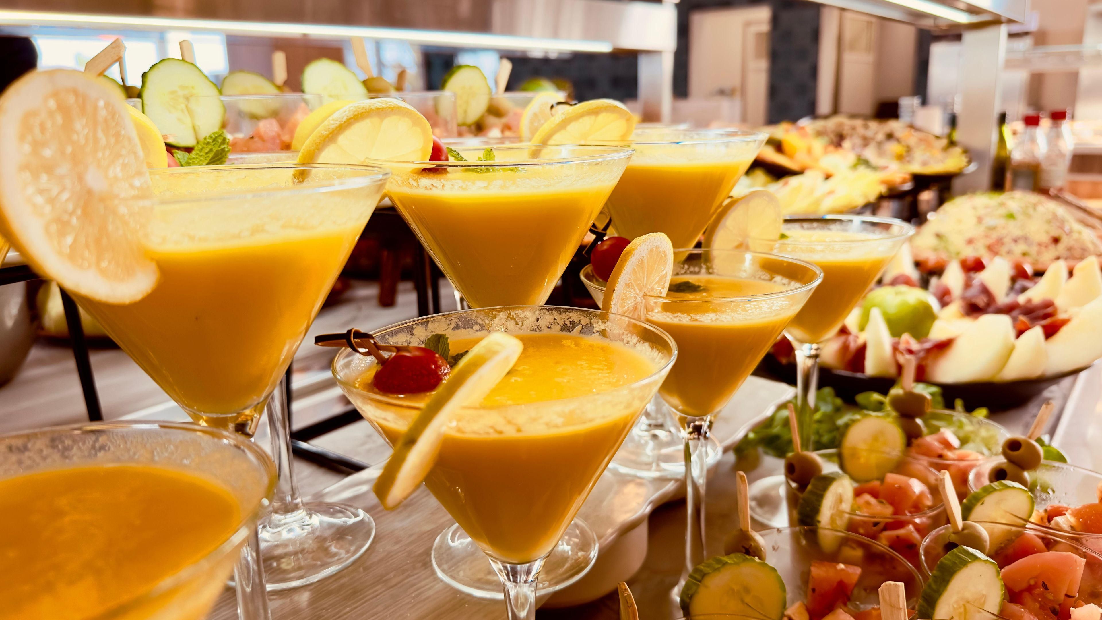 A buffet with various food items and a selection of orange-colored drinks in martini glasses.