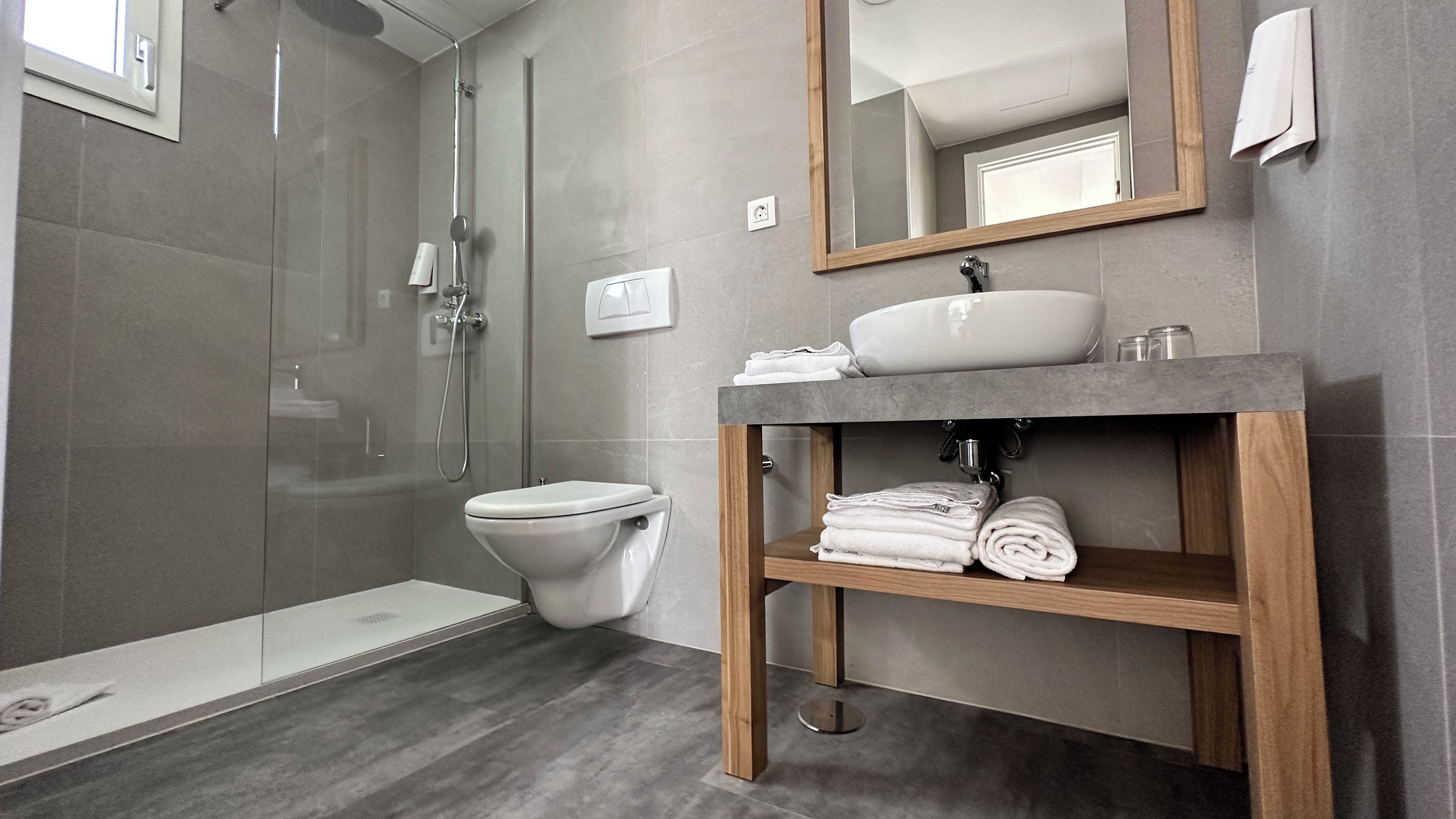 Ein modernes Badezimmer mit einer ein Glasfach raumseitig abgeschirmten Dusche, einer Toilette, einem Waschbecken mit Holzschublade und einem darüber hängenden Spiegel.