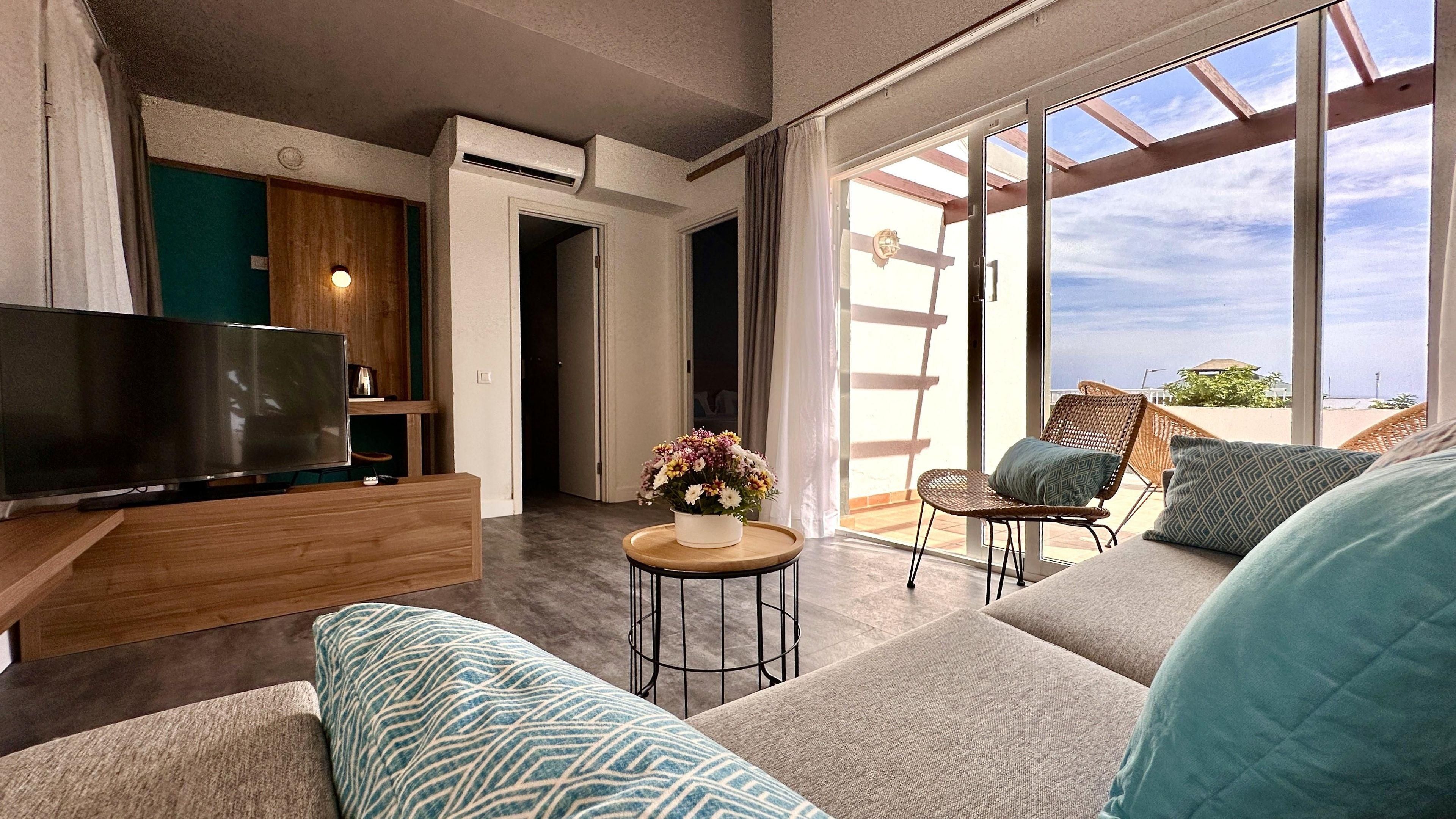 Una sala moderna con una televisión de pantalla plana, un sofá, una mesa de centro con un arreglo floral y un balcón con mobiliario de mimbre.