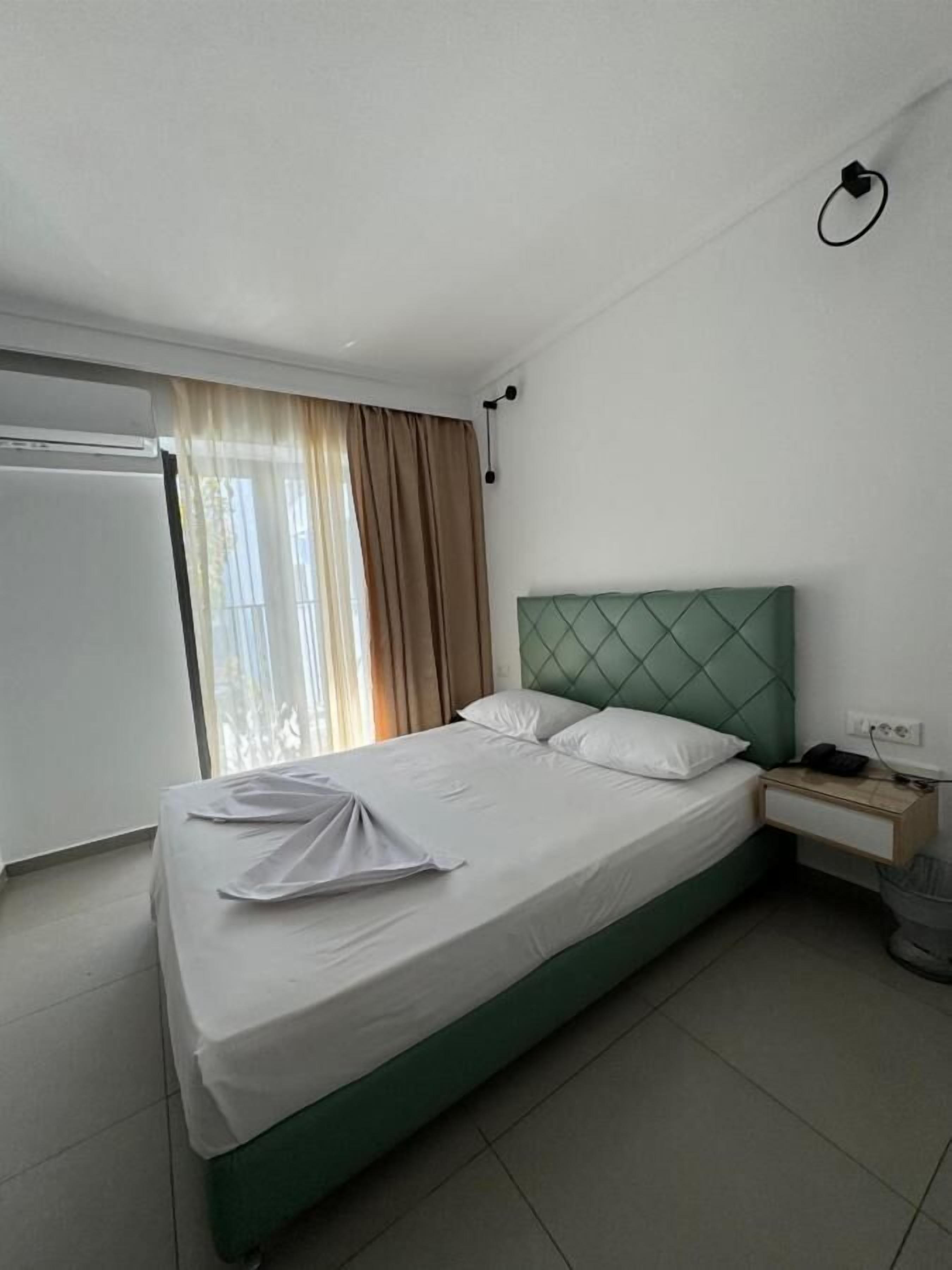 Quarto de hotel com uma cama, mesa de cabeceira, abajur e janela com cortinas.